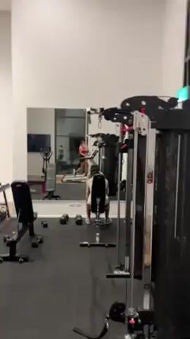 The scene unfolds in a well-equipped gym. In the background, a woman in a workout outfit is on a treadmill. A man is seated on a weight machine with his back to the camera. Various pieces of gym equipment, including a bench and dumbbells, are scattered throughout the room. A large mirror reflects the gym's activities, while natural light enters through a tall window. The gym appears to be clean and organized.
