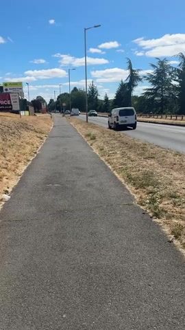 This media file captures a sunny day in Reading, United Kingdom, with a clear blue sky dotted with white clouds. The scene is a roadside, with a paved pedestrian footpath running parallel to a busy road. On the left side of the footpath, there is dry, brown grass and some vegetation, leading up to a few signs, one of which prominently displays "ABBEY Roofing Trade" and "DON'T BE BEAT ON PRICE." Further down the road, more signs and buildings are visible.

The road itself has a few cars and white vans driving on it, indicating moderate traffic. Tall streetlights line the footpath. On the right side of the road, there are trees, including some mature conifer trees, and a fence, suggesting a boundary to a property or area beyond the road. The overall impression is of a suburban or semi-rural setting on a warm, bright day, likely in the summer given the dry grass and clear skies. No specific activity or event is discernible beyond the general movement of traffic and the presence of pedestrians on the footpath, who are too distant to be identified. The time of day appears to be midday or early afternoon due to the bright sunlight and the high angle of the shadows.