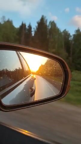 The video shows a road at sunset as seen from the passenger side of a car. The sun is low in the sky, and the light is reflecting off the wet pavement of the road. The side mirror of the car is visible, with water droplets on the glass, and the sun is reflected in the mirror. Trees line the side of the road. The car is moving on the road. The sky is a light orange color. The video focuses on the reflection and light on the mirror and window of the car. There is music playing.