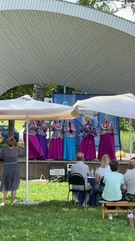 A group of five women in matching colorful outfits and long skirts are performing on a raised wooden stage under a white canopy. They are all singing and gesturing with their hands. One woman in a blue dress is in the center, while the others are in purple dresses. Behind them is a blue banner with text and graphics. In front of the stage, there is an audience of several people sitting on chairs and benches. The area is outdoors, with grass and trees visible. There are also two large white umbrellas in the foreground, partially obscuring the view of the stage. A woman in a dark dress is standing in the foreground facing the stage.