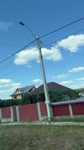A camera pans across a suburban area under a clear blue sky with scattered white clouds. The scene includes houses with red roofs and fences. A large blue truck is parked on the right side of the frame, followed by a white semi-truck with an orange trailer. Power lines are visible crisscrossing the sky. The camera moves past a small tree and a telephone pole. The video ends with a view of more houses and trees.