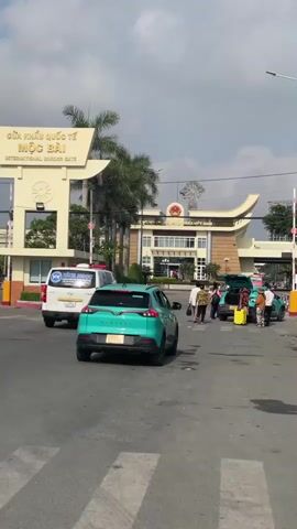 The video depicts a border crossing at the Mộc Bài International Border Gate. Several people and cars are present, moving through the crossing. The architecture of the border gate is visible, and the sky is partly cloudy. There are palm trees and other greenery dotting the landscape. In particular, there are two turquoise Vinfast VF e34 vehicles driving on the road. One of the cars has an open trunk and several people standing near it. The scene appears to show normal border crossing activity.