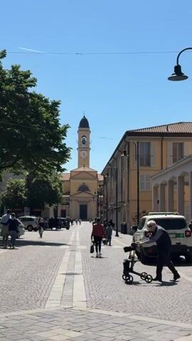 The video depicts a street scene in what appears to be a European town. The street is paved with cobblestones and lined with buildings. In the distance, a church with a tall clock tower is visible. Several people are walking along the street. A person with white hair is walking towards the church using a wheeled walker. A few parked cars, including police vehicles, are visible along the sides of the street. The sky is clear and blue.
