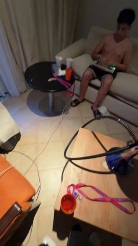 A shirtless person is sitting on a light-colored couch wearing black shorts with white stripes and black sandals.  They are operating a small electronic device, possibly a DJ controller, that sits on their lap. The person has tattoos of a cross and the number "1999" on their lower right leg. The scene includes a black round table with disposable cups, an orange chair, a hookah on a wooden table, and various cables on the floor.  The room has tiled floors and light-colored walls.  The music is loud and has a dance beat.
