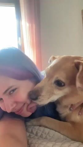 A woman with purple hair, lying on a bed, is being licked on the face by a light brown dog. The woman is smiling and holding the dog. A window and a framed picture are visible in the background.