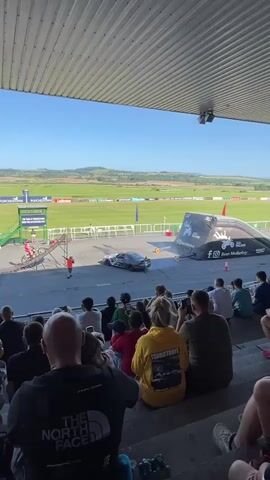This is a low-angle, medium shot video of a stunt performance. In the foreground, an audience sits in tiered seating, facing away from the camera. Their backs are to the viewer, and some are holding up phones to record. In the midground, a motorcyclist in a red suit performs a jump over a silver car. A large inflatable ramp is positioned to the right of the car, with the motorcyclist airborne above it. To the left of the car is a smaller ramp, and a crew member in an orange shirt stands near it. The background shows a wide, green field with a clear blue sky. The scene appears to be outdoors, possibly at a sporting event or festival.