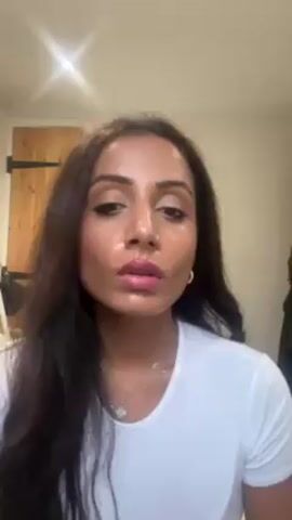 A woman with long brown hair, a nose ring, gold hoop earrings, and a silver necklace is putting on mascara in a room with a white wall, a wooden door with black metal hardware, and a lamp in the background. She is wearing a white T-shirt. She is looking up as she applies mascara to her upper eyelashes. She then pauses and looks down at the mascara wand.
