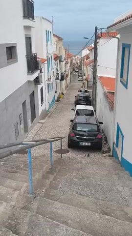 The video shows a steep, narrow cobblestone street lined with white buildings. Several cars are parked along the street, and there are stairs leading down. The buildings have red-tiled roofs and blue trim around the windows. In the distance, the ocean is visible. The sky is overcast.