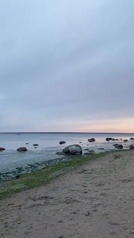 The video shows a beach with rocks and water. The sky is cloudy and there is a sunset. There are two people standing on the beach in the distance. One person is holding a camera and taking a picture of the other person.
