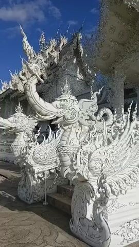 The video shows a close-up panning shot of an ornate white temple, likely a Buddhist temple, under a blue sky with some clouds. The architecture is highly detailed and intricate, featuring white sculptures of mythical creatures, possibly dragons or serpents, adorning the steps and the facade. The structures are characterized by elaborate carvings, pointed spires, and a shimmering, almost crystalline appearance, suggesting the use of reflective materials like glass or mirrors. The camera movement is slow and deliberate, focusing on the detailed craftsmanship and the overall grandeur of the temple. There are no people clearly visible in the frame, and the environment appears to be an outdoor setting. The lighting suggests it is daytime, with sunlight reflecting off the white surfaces.