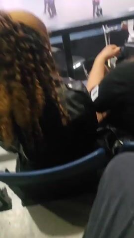 The video shows an indoor arena filled with fog, with the camera positioned behind people in a seated area. In the distance, people dressed in purple and yellow uniforms stand around a large, open area. Some of the seated viewers are looking at their phones. The lighting is bright, with spotlights shining down. The camera angle is slightly elevated, looking down onto the scene.