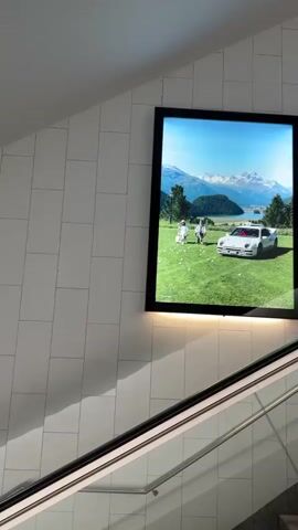 A framed image is displayed on a white tiled wall above an escalator. The image depicts a white sports car parked on a grassy field with a lake and mountains in the background. There are several people in the image, appearing to be enjoying the scenery. The lighting in the video suggests it is daytime, with sunlight illuminating the scene. The escalator is visible in the foreground, moving upwards. The video is shot from a low angle, looking up at the artwork.