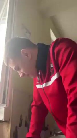 The video shows a young man wearing a red tracksuit with white and black accents. He is standing indoors in a room with sunlight streaming in from a window on the left. The man is looking down initially, then looks up and smiles. The walls are painted a neutral color, and there are some posters or pictures on the wall behind him. The man's expression changes from neutral to smiling, and he appears to be in a hallway of some kind.