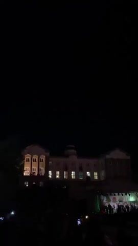 At night, a crowd of people face a stage in front of a lit building. Fireworks explode in the sky. The camera is hand-held.