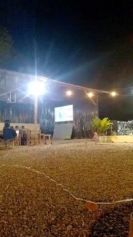 The video shows an outdoor area at night. The ground is covered in gravel. In the background, there is a covered structure with corrugated metal roofing and bamboo-like walls. A white screen is visible on the wall of the structure. Several people are seated on chairs in front of the screen. There are string lights hanging above. The scene is lit by artificial lights, creating bright spots and shadows.