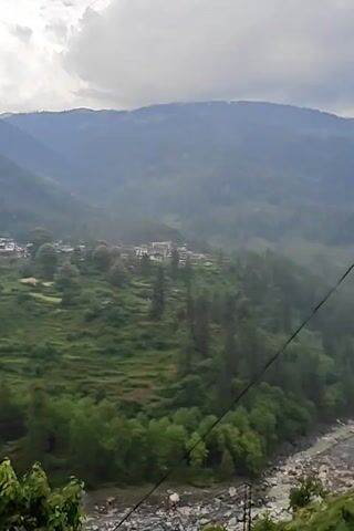The video presents a scenic mountainous landscape under an overcast sky. In the foreground, green foliage from an unseen plant is visible at the bottom left. Below this, a rocky riverbed with a flowing river occupies the lower portion of the frame. Across the river, a lush green valley unfolds, characterized by terraced fields and dense tree cover, including tall pines and other deciduous trees. Nestled within this valley is a village or small town, identifiable by several buildings, some featuring prominent red roofs. Towering forested mountains form the backdrop, their peaks partially shrouded in clouds. The overall impression is one of a serene, natural environment with human settlement integrated into the landscape. The camera slowly pans slightly to the right throughout the clip, offering a broader view of the vista. No specific human activity is visible, and no text is present in the video.