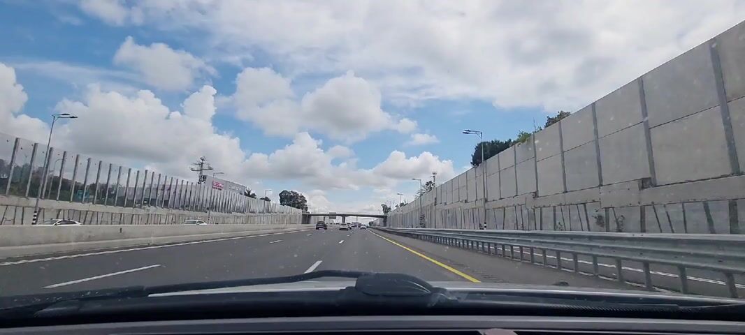 The video shows a car driving down a highway on a partially cloudy day. The road has multiple lanes with yellow and white lane markings. A concrete wall lines the right side of the highway, while the left side is bordered by a fence and buildings in the distance. Overhead, a bridge spans the highway. Other cars are visible in the distance.
