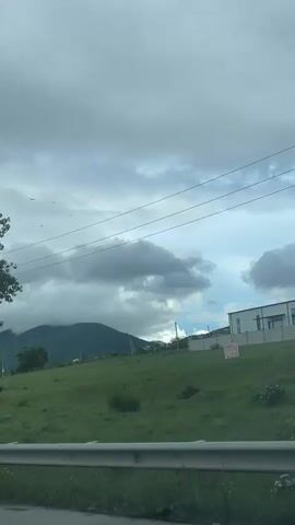 The camera pans from a view of a tree to a landscape showing a mountain partially obscured by low clouds on a cloudy day. The foreground includes roadside vegetation and a guardrail. Power lines and poles run across the sky. As the camera continues moving, it passes under a roadside sign structure. The video is shot from a moving vehicle on a road.