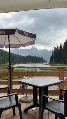 This media file captures a scenic view from a covered outdoor seating area, likely a restaurant or cafe, in Auronzo di Cadore, Italy. The foreground features a wooden deck with a black plastic table and two chairs. An open umbrella, adorned with the logo "NUII", is positioned to the left of the table, offering shade.

Beyond the deck, a picturesque landscape unfolds. A wooden railing separates the deck from a natural setting that includes tall green pine trees bordering a tranquil lake. In the distance, a majestic mountain range rises, its peaks partially obscured by a cloudy sky. The overall impression is one of a serene, natural environment, possibly experienced during the daytime given the lighting.

The weather appears to be overcast, with significant cloud cover, contributing to a diffused light. There are no people visible in the frame, suggesting a moment of quiet observation or perhaps the area is temporarily unoccupied. The setting combines man-made elements for comfort and relaxation with the raw beauty of the Italian Alps and a clear lake. The visible text "NUII" on the umbrella is the only prominent text in the media.