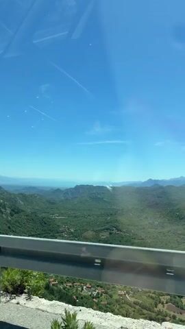 This is a vertical, shaky video shot from a moving vehicle, looking out over a vast, lush green landscape of rolling hills and mountains. The sky is a clear, bright blue with a few faint contrails from airplanes visible. In the foreground, a silver guardrail runs along the edge of the road, with some dry grass and small plants growing by its base. Beyond the guardrail, the landscape stretches out, with dense vegetation covering the hills. In the distance, the mountains become hazy, and a body of water, possibly a lake or the sea, can be seen on the horizon. The overall impression is one of natural beauty and a sense of open space.