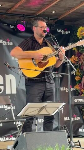 A man stands on a wooden stage, playing an acoustic guitar and singing into a microphone. He is wearing a black striped short-sleeved shirt and black pants. The stage is set up outdoors, with a wooden ceiling overhead. Behind him, a black banner with pink text that reads "Bakgården" is visible, along with some floral decorations and a Yamaha keyboard. The lighting includes pink stage lights. A music stand with sheet music is positioned in front of the man.