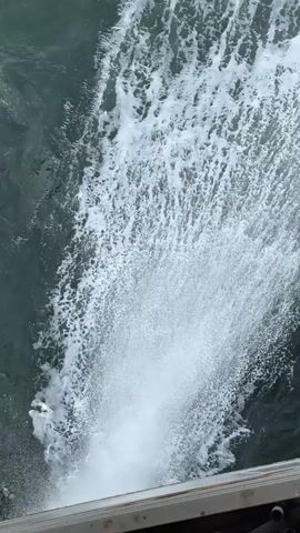 From an elevated position, a high-angle shot depicts a torrent of white, foamy water cascading into darker, calmer waters. The cascading water creates a wide, frothy spray upon impact, contrasting with the surrounding deeper, greenish-blue sea. A sliver of what appears to be a gray or metal structure is visible at the bottom of the frame, suggesting the vantage point is from a bridge or platform. The scene captures the dynamics of water movement and the visual impact of a concentrated water discharge into a larger body of water.