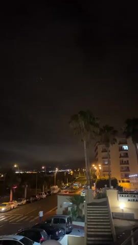 The video shows a street scene at night. The sky is dark with some clouds and a few faint lights that may be stars or distant lights. A street with cars parked along the side and some cars driving is visible. Palm trees are also present. On the right side of the frame, there is a building with lights on, and a set of stairs leading up to it. A sign with text is visible on the building. The overall atmosphere is that of a quiet urban night.