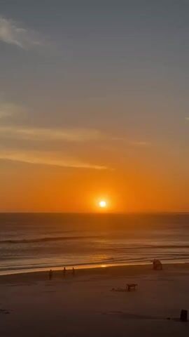 The video shows a sunset over the ocean. The sun is a bright orange ball on the horizon. The sky is a gradient of orange, pink, and blue. The ocean is calm with small waves. There is a beach in the foreground with a few people walking and sitting. The overall mood is peaceful and serene.