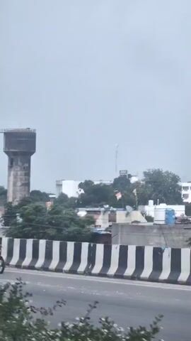 The video shows a water tower in the background, a street light in the foreground, and vehicles driving on a road. The sky is overcast. The water tower is a tall, cylindrical structure with a square base and a cylindrical top. It appears to be made of concrete. The street light is a tall, slender pole with a light fixture at the top. The vehicles are cars and a construction vehicle. The road is a multi-lane highway with a black and white barrier separating the lanes. The environment appears to be an urban or suburban area, with buildings and trees visible in the background.