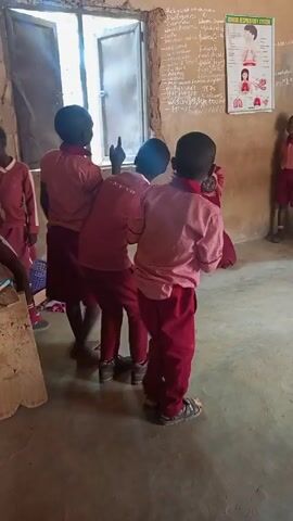 The video shows a classroom scene with several children dressed in maroon and pink uniforms. Three children are standing close together, facing a chalkboard covered in writing, and gesturing with their hands and arms in what seems like a coordinated movement or dance. Another child stands to the side, near the chalkboard, watching them. There is a poster of the human respiratory system on the wall. The classroom appears simple, with a concrete floor and a window providing natural light. Additional children are visible in the background, sitting at desks.