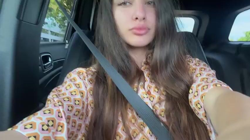 The video shows a young woman in the driver's seat of a car, wearing a patterned shirt and a seatbelt. She is looking at the camera and appears to be taking a selfie or recording a video. The interior of the car is visible, including the dashboard and seat. Sunlight is filtering in from outside, casting a warm glow on the scene. The woman's hair is long and dark, and her eyes are green. Her lips are slightly pursed. The overall impression is a casual, personal moment captured while in transit.