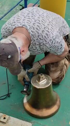 A man kneeling on a green surface is using a power tool to polish a ship's bell. The bell, which is partially polished, is shaped like a traditional nautical bell, with the upper portion being shiny silver and the lower portion a dull copper color. The man, wearing a baseball cap, gloves, and a T-shirt with a fish pattern, is focused on the polishing process. His posture suggests he is applying steady pressure to the tool as he works. The background includes a yellow cylindrical object and blue metal railings, suggesting the activity is taking place on a boat or dock.