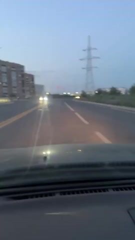 The video shows a view from inside a vehicle, looking out the windshield at a road at dusk. The sky is a pale blue, transitioning to darker hues. On the left side of the road, there are several tall, multi-story buildings. Further down the road, on the right, is a large electricity pylon with wires extending across the sky. The road itself has visible lane markings, including a double yellow line on the left and a white line on the right. Headlights from other vehicles are visible in the distance, and streetlights are also present. The dashboard of the car is visible in the foreground. The audio consists of ambient sounds and spoken words in Russian. The speaker expresses that they have driven somewhere "not there" and that they should have turned around.