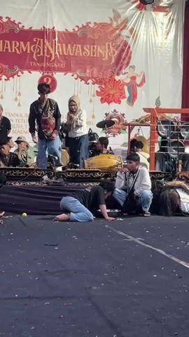 A group of people are gathered on a stage in front of a banner. Two individuals, a male and a female wearing a headscarf, stand center stage. The male is wearing traditional Indonesian clothing, including a batik shirt and sarong, and appears to be performing or posing. The female is wearing jeans and a t-shirt. To the right of the stage, there is a musical ensemble with instruments, including gongs and drums. Several other people are seated and standing around the stage, some observing the performance. In the foreground, a male wearing a black shirt and jeans is lying on the floor, his body curled up. Another male, wearing a white shirt and jeans, is sitting to his right, looking towards the stage. The floor is covered in a dark material, possibly a carpet or tarp, with some debris scattered on it. The overall environment appears to be an indoor venue, possibly a community hall or cultural center, with lighting fixtures visible above the banner.