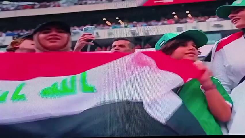The video captures a crowd of spectators and participants in a sporting event, likely a soccer match given the presence of flags and player attire. In the foreground, a young boy holds up an Iraqi flag. Behind him, several individuals are visible, including a man in a white jacket with the Iraqi flag emblem, and another man in a blue vest with "VOLUNTEER" printed on the back. Spectators are seated in the background. The overall atmosphere appears to be one of anticipation and national pride.