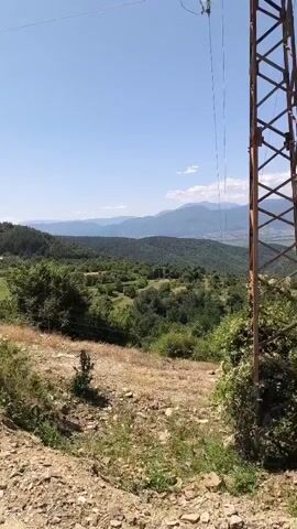 A handheld video pans across a mountainous landscape on a clear, sunny day. A tall, metal power line tower stands in the foreground, partially overgrown with green foliage. Beyond the tower, rolling hills covered in lush green trees stretch into the distance. In the far background, a range of hazy, blue mountains forms the horizon. The ground in the foreground is dry and rocky, with sparse patches of grass. The sky is a vibrant, cloudless blue, with a few wispy white clouds visible near the horizon. The camera movement is smooth and deliberate, capturing the expansive vista.