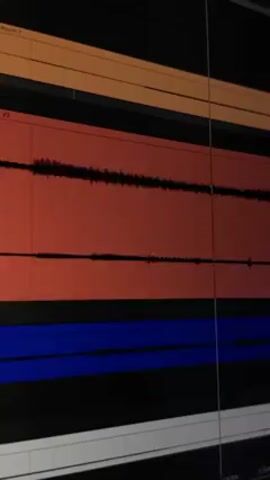 A close-up shot shows a computer screen displaying an audio editing interface. The software presents multiple audio tracks distinguished by different colors: orange, red, and blue. The waveforms of the audio signal are visible within each track. The screen is lit with the glow from the software.
