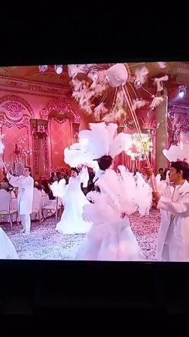 This video captures two distinct scenes. The first scene, appearing at the beginning, shows a lively indoor event, possibly a wedding reception. Several individuals are dressed in white, appearing to be performers or participants in a celebration. They are engaged in an activity that involves large, feathery white props, creating a visually striking effect with plumes of white material being tossed or unfurled. The environment seems ornate, with pink-toned lighting and decorative elements suggesting a grand venue.

The second scene, which begins around the 00:02 mark, shifts to a different setting, likely a kitchen or food preparation area. A person wearing a white chef's coat is actively stirring a large quantity of ground meat in a massive, dark, round cooking vessel. The food is visibly being cooked, with steam rising from the vessel. The cooking surface appears to be a large, flat, black griddle or pan set over a heat source. The surrounding environment shows industrial kitchen elements, including a tiled floor and a metal chimney or exhaust pipe. The focus is on the culinary process and the chef's action of stirring the ingredients.