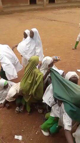 In this video, a group of children, predominantly girls wearing white hijabs and green pants, are gathered in an outdoor, dirt-floored area. The scene appears to be a schoolyard, with a building visible in the background. The children are engaged in a playful tug-of-war with a green cloth. Some children are on the ground, holding onto the cloth, while others are standing and pulling. The atmosphere seems energetic and joyous, with the children appearing excited and focused on the game. The time of day is indeterminate, but the lighting suggests it could be daytime. There is no visible text in the video. The location context is Kagarko North, Nigeria.