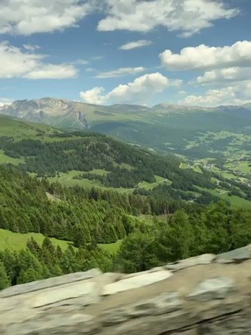 The video shows a panoramic view of a lush green valley with mountains in the background. The sky is blue with scattered white clouds. In the foreground, there is a stone wall. The camera is moving, giving the impression of a car or train journey. The valley floor is dotted with small buildings and appears to be farmland. The mountainsides are covered in dense evergreen trees, with some patches of lighter green indicating meadows or sparser vegetation. The overall scene conveys a sense of natural beauty and tranquility.