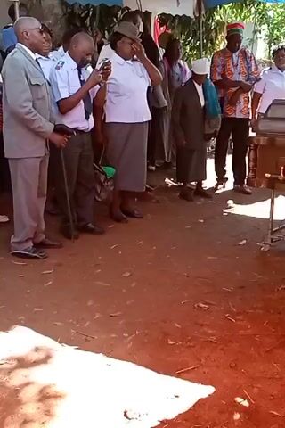 Here is a description of the video.

The video shows a group of people gathered outside in what appears to be a funeral setting. A casket is visible, surrounded by people who appear to be mourning. The environment is outdoors with red dirt on the ground, possibly in a rural area. 

The individuals present are dressed in a variety of clothing. Some are wearing uniforms, while others are in casual attire. One person is holding a cane, and another is using a mobile phone, potentially to record the event. The overall atmosphere is somber, as one would expect at a funeral.