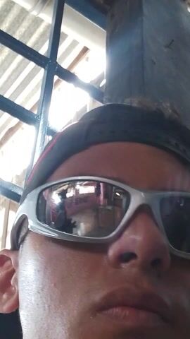 The video shows a close-up shot of a person wearing reflective sunglasses and a black headband. The person's face is sweaty. The glasses reflect the interior of a room. Above the person, a window with a blue-painted frame and metal bars can be seen. The background behind the window is bright, suggesting daylight. A song plays in the background.