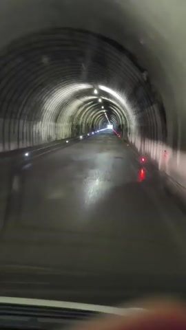 The video shows a point-of-view perspective of traveling through a long, dimly lit tunnel. The tunnel has a curved ceiling with visible supporting arches and overhead lighting. The road surface appears wet, reflecting the lights. Red and white lights are visible along the sides of the tunnel, marking its edges and path. The tunnel extends into the distance, with a brighter light visible at the far end, suggesting the exit. The lower portion of the video shows the dashboard and windshield of a vehicle.