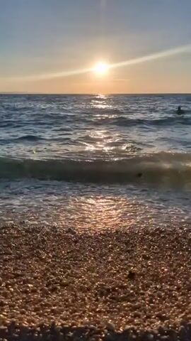 This is a short video clip of a beach scene in Zadar, Croatia, likely taken at sunset. The sun is low on the horizon, casting a warm, golden light across the water and the pebbled shore. Gentle waves are rolling in, creating a soft foam as they reach the beach. In the distance, a person can be seen swimming or wading in the sea. The overall mood is peaceful and serene, capturing the beauty of a coastal evening.