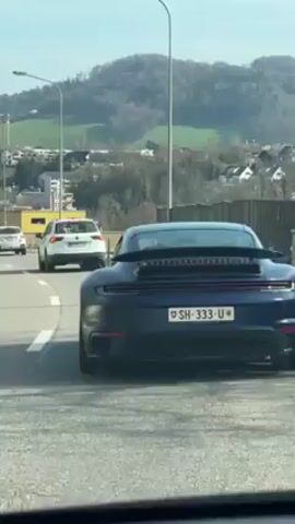 The video shows the back of a dark blue Porsche 911 Turbo S car with a Swiss license plate (SH-333-U) as it is driving on a highway in Switzerland on a sunny day. The car is in a line of traffic and is moving slowly. In the background, there are green hills, trees, buildings, and street lights. There is also a white Volkswagen car driving behind the Porsche.