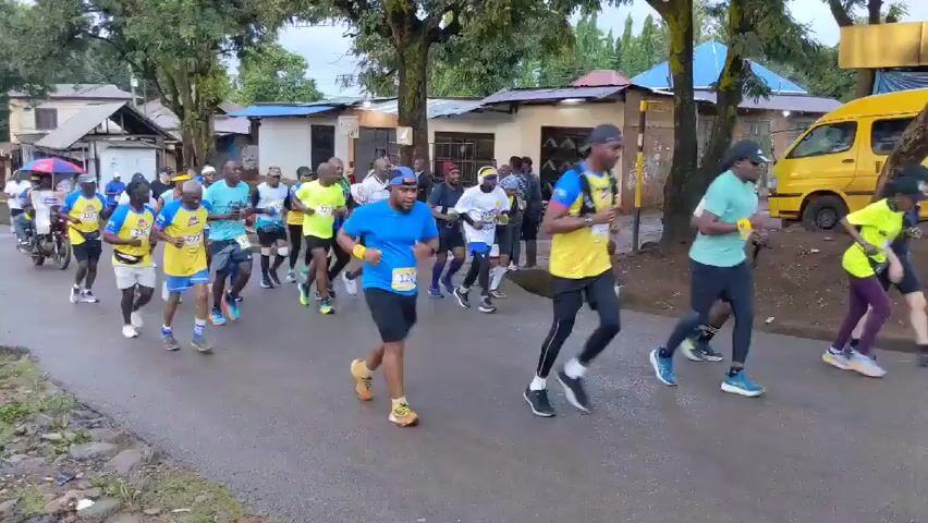 A group of about 20 people, mostly men, are running down a paved road in what appears to be a race. They are wearing athletic clothing and race bibs. Several are wearing yellow and blue jerseys. Some wear shorts, while others wear long leggings. The road is lined with trees and small buildings. Several yellow vans are parked on the side of the road. A motorcycle with two people on it follows the runners. People are shouting encouragement to the runners in an unidentified language. It appears to be overcast.