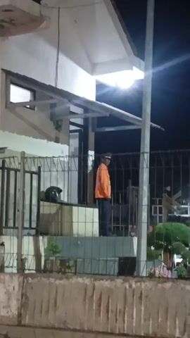 A person wearing an orange vest and a dark hat stands on a raised platform in front of a building at night. A fence runs in front of the platform, and a motorcycle helmet rests on the platform to the left of the person. There is a light fixture illuminating the area.