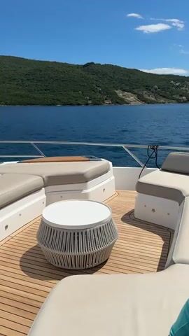 The video shows the bow of a modern yacht moving through calm blue water under a clear blue sky with some clouds. The yacht has a wooden deck and white seating areas. A small white table with a woven design sits on the deck. In the background, a lush green, tree-covered mountain range slopes down to the water. A small purple flag with a crest is visible at the front of the yacht. The camera pans across the scene, offering views of the yacht's exterior and the surrounding landscape.