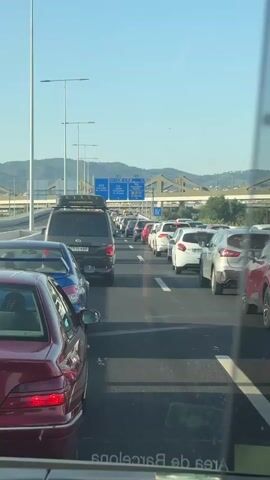The video shows a long line of cars stuck in traffic on a highway. The sun is shining, and the sky is clear blue. The cars are all facing forward, and some of them are very close together. The highway has multiple lanes, and there are large blue signs overhead indicating directions to various cities, including Barcelona and Girona. There are also mountains in the background. The camera appears to be inside one of the cars, looking forward through the windshield. The video is shot from a slight upward angle.