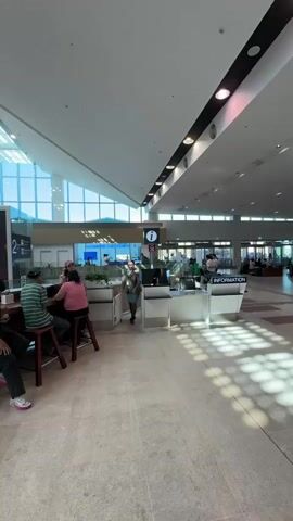 The video shows a busy airport terminal. In the foreground, people are seated at a bar. There is a large screen displaying text and images. A luggage cart is visible. The terminal has a high, vaulted ceiling with skylights and lighting fixtures. In the background, there is an information desk with staff members. Further in the distance, there are check-in counters and lines of people. People are walking with luggage. The floor is tiled, and there are patterned inlays. The overall environment appears to be a modern, well-lit airport.